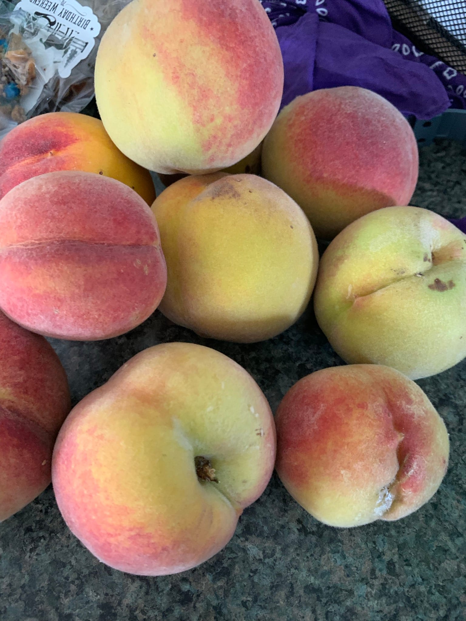 A group of plump, fuzzy peaches is piled on a slate grey and black stone countertop.