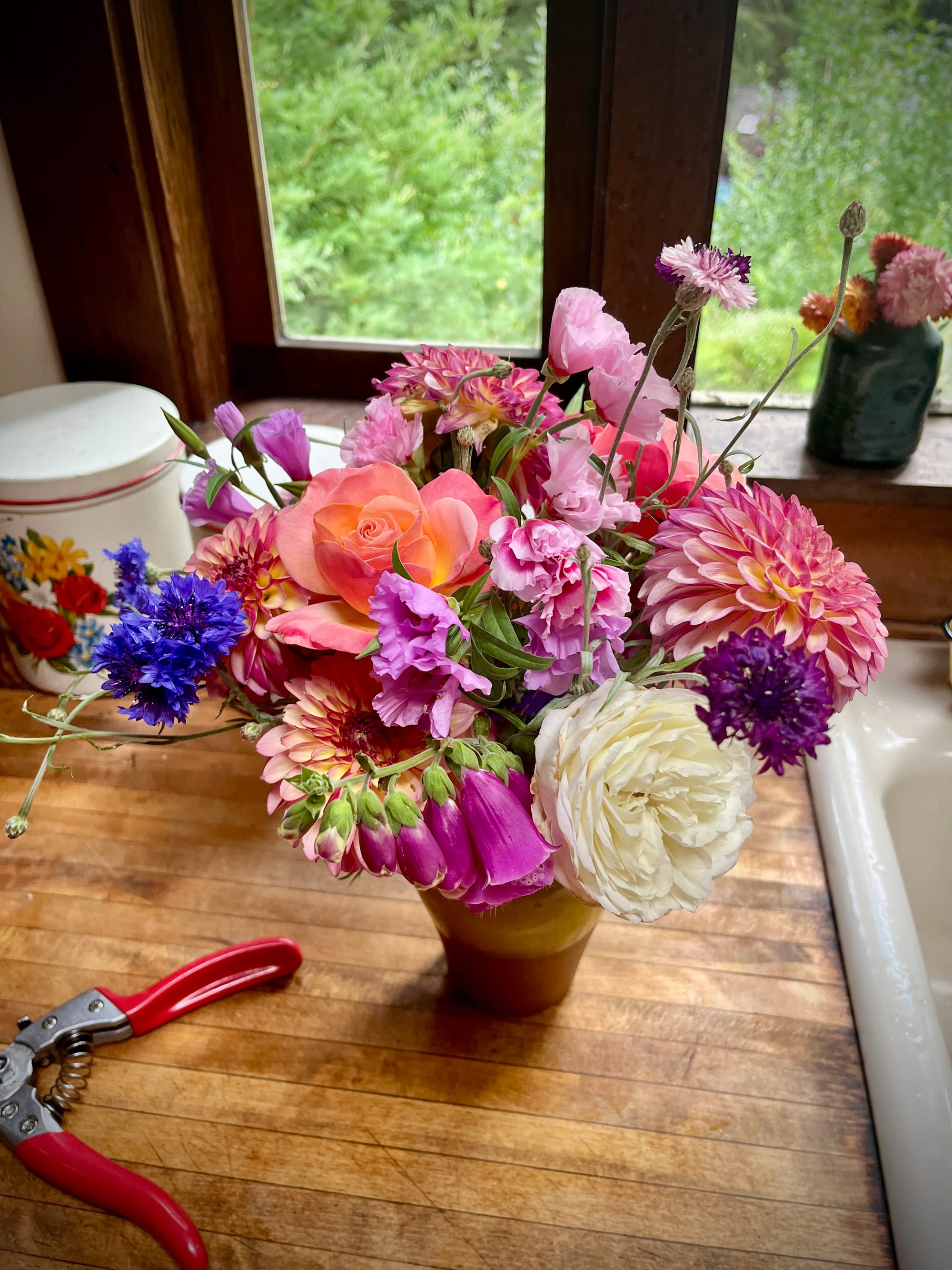 Mustard yellow vase filled with white, purple, rose, and cornflower blue flowers sitting on a wooden countertop, with a pair of red handled floral trimmers barely in frame