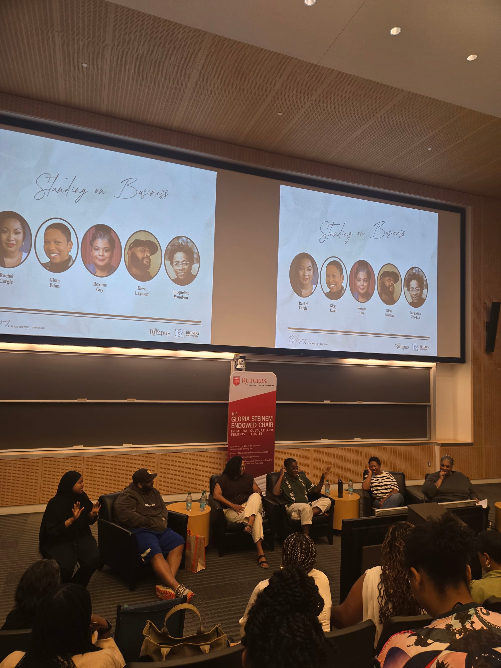 Image of six individuals seated in front of a chalkboard in front of a crowd.