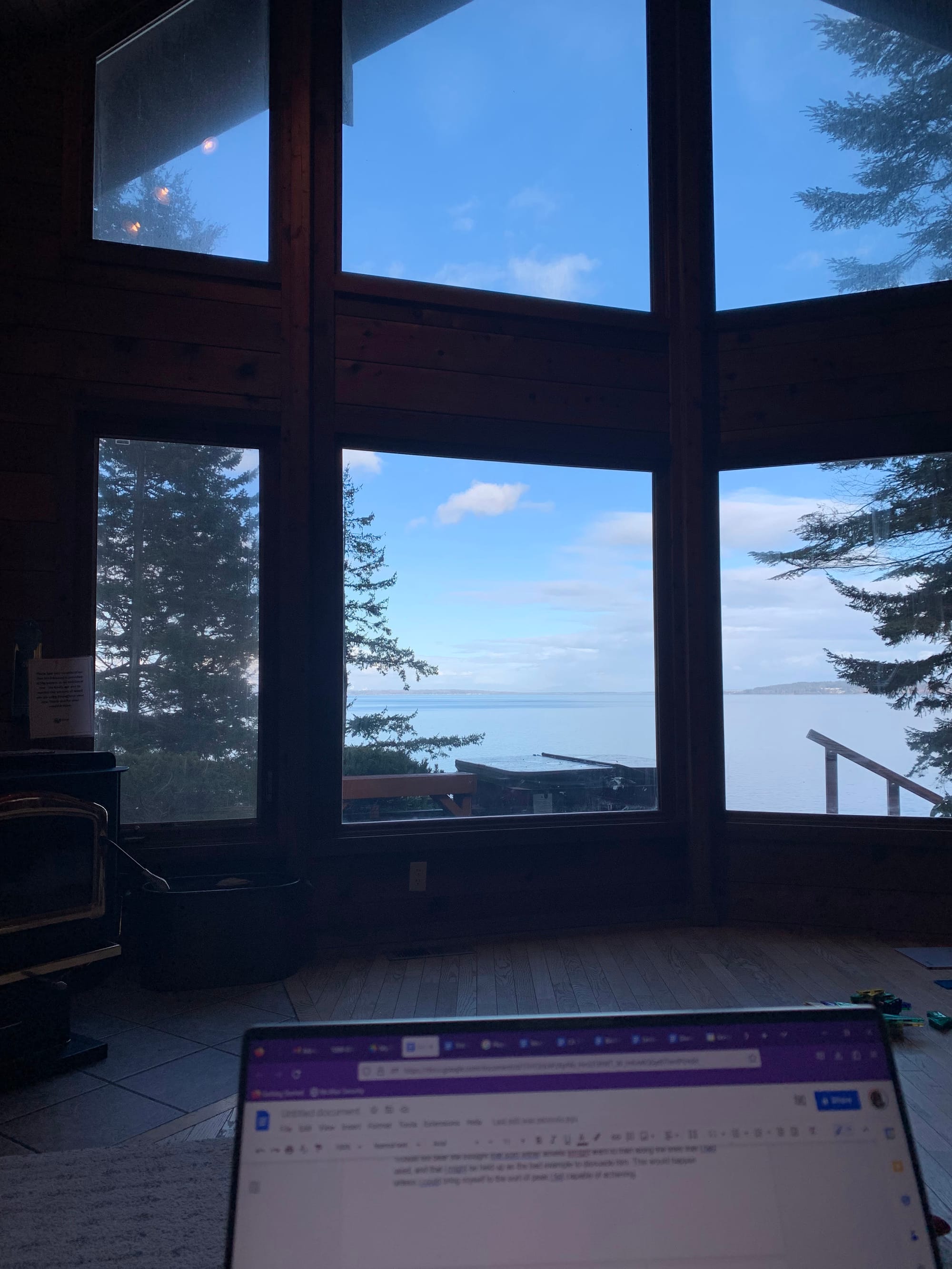 six large windows look out on blue skies and trees and water, in the foreground, a laptop is open