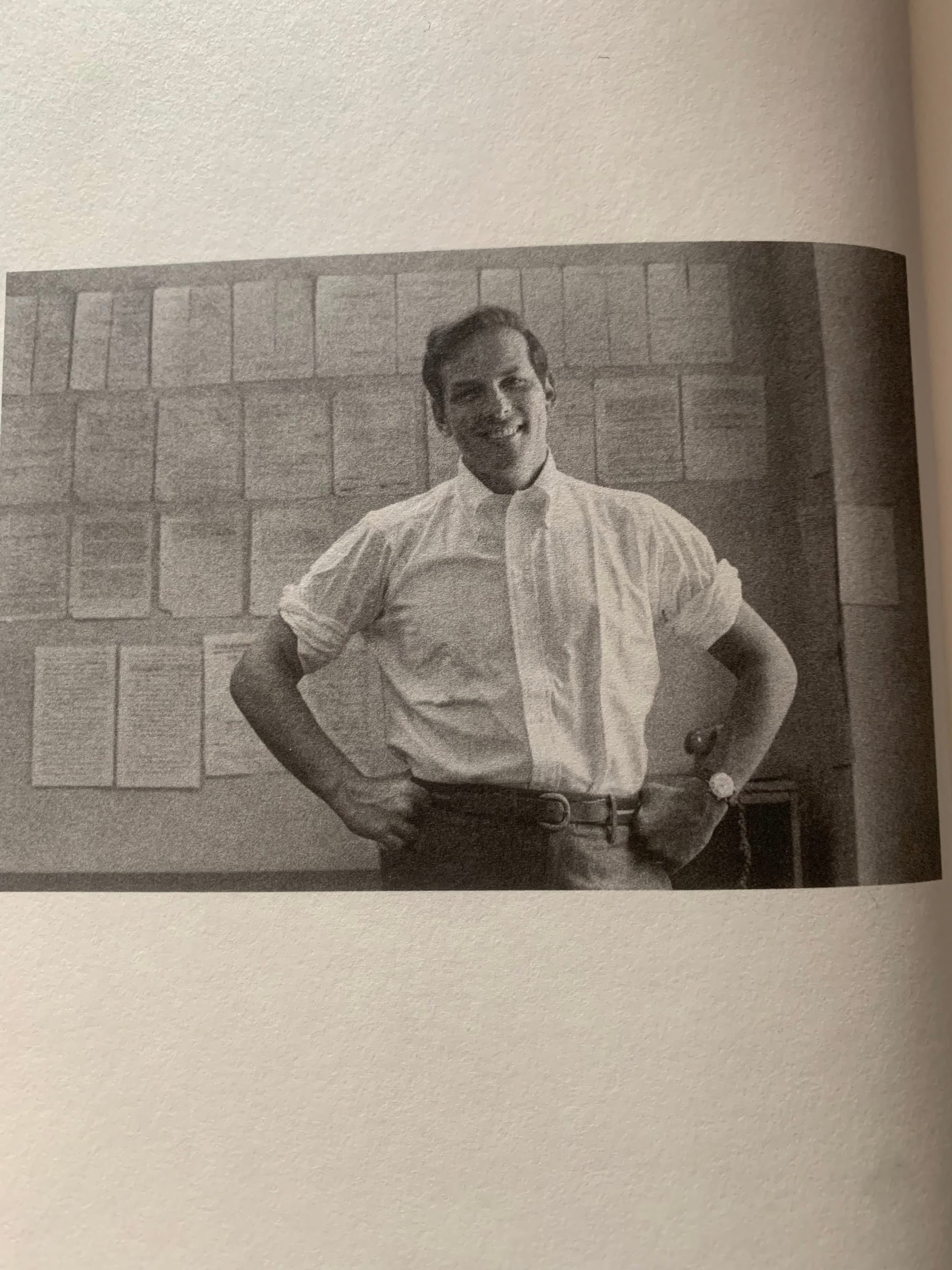 A black and white photo on a book page, of a white man in front of papers pinned to the wall. He is wearing a white collared shirt with rolled up sleeves and smiling.