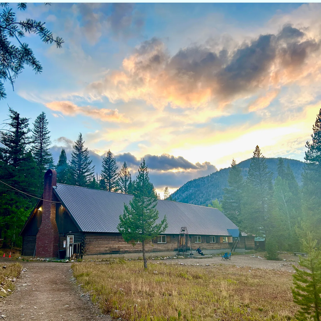 A long brown log cabin with brick chimney sits in a large field with a mountain and tall trees in the background.