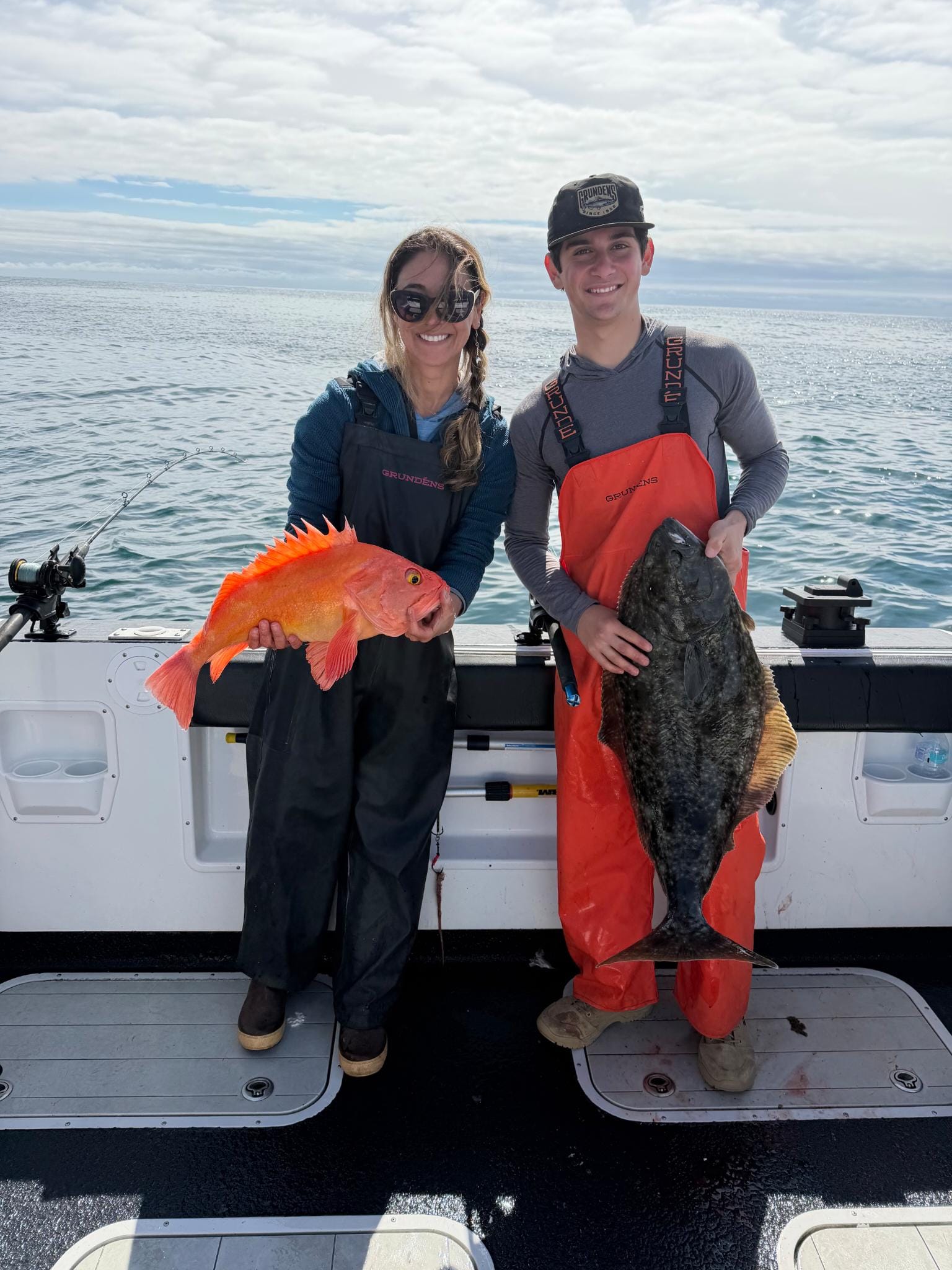 A woman with her hair in a braid and fisherman waders on holds a huge red fish, next to her a young man in a hat and orange waders holds a large grey fish. They are on a boat with open water behind them.