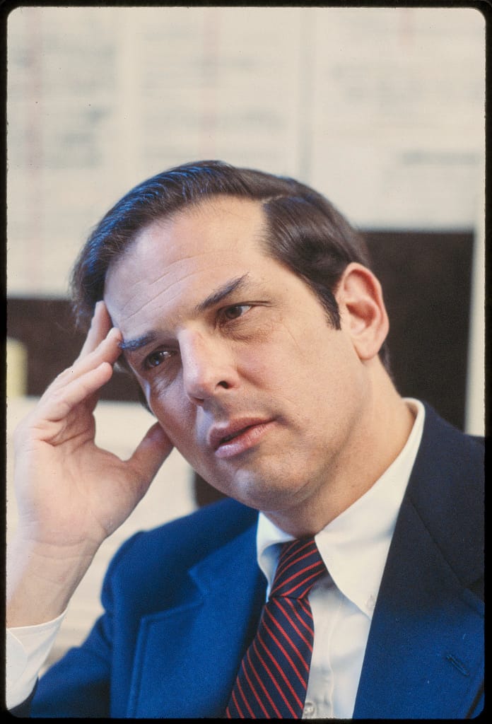 A portrait of a white man with brown hair in a blue suit and red and blue tie, he rests his head on his hand.