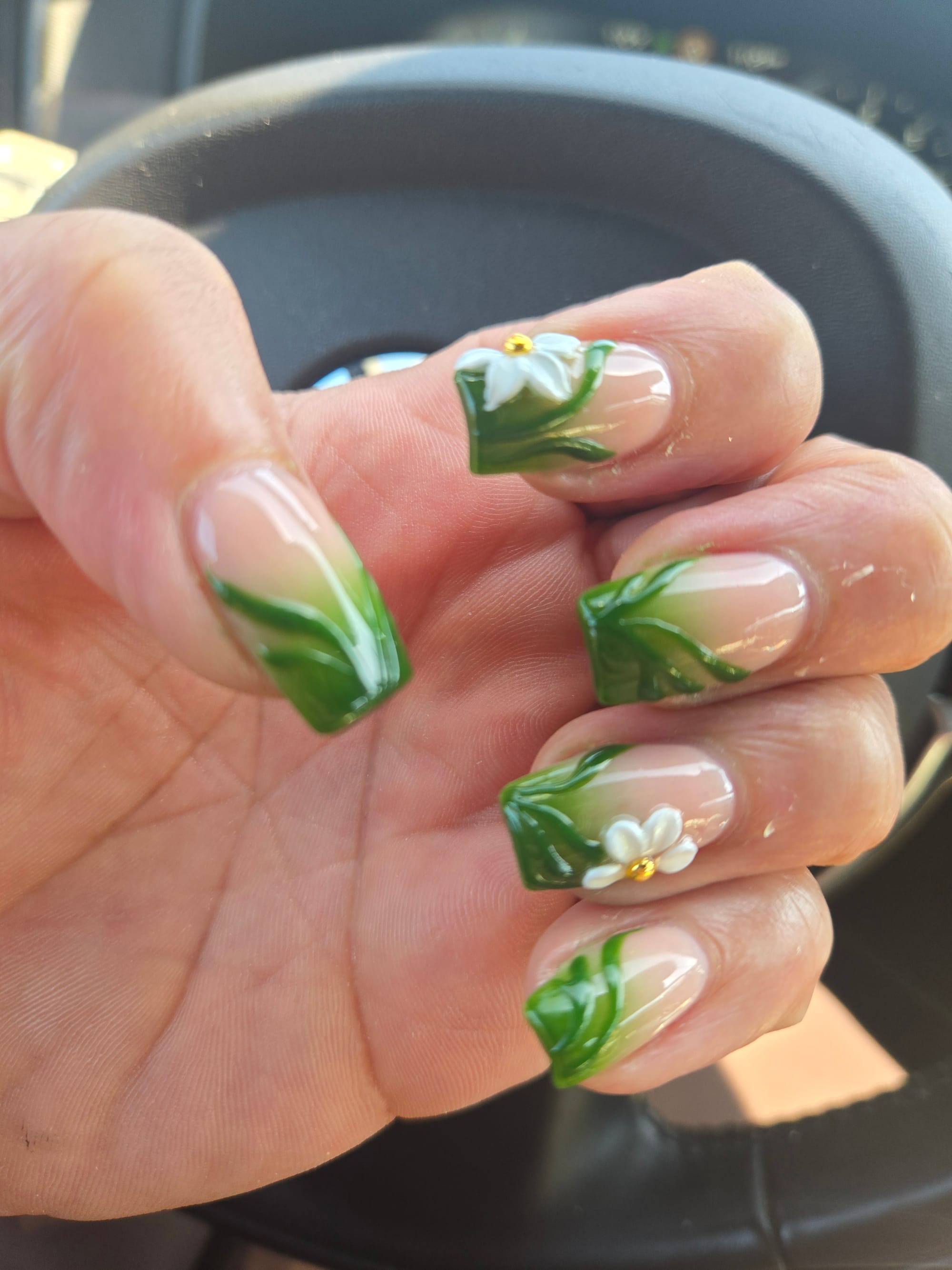 Close up of a person's left hand featuring medium length square nails with a green French manicure. Green vines crisscross the nails, and 3D white flower decorations with small gold bead centers grace the index and ring fingers.