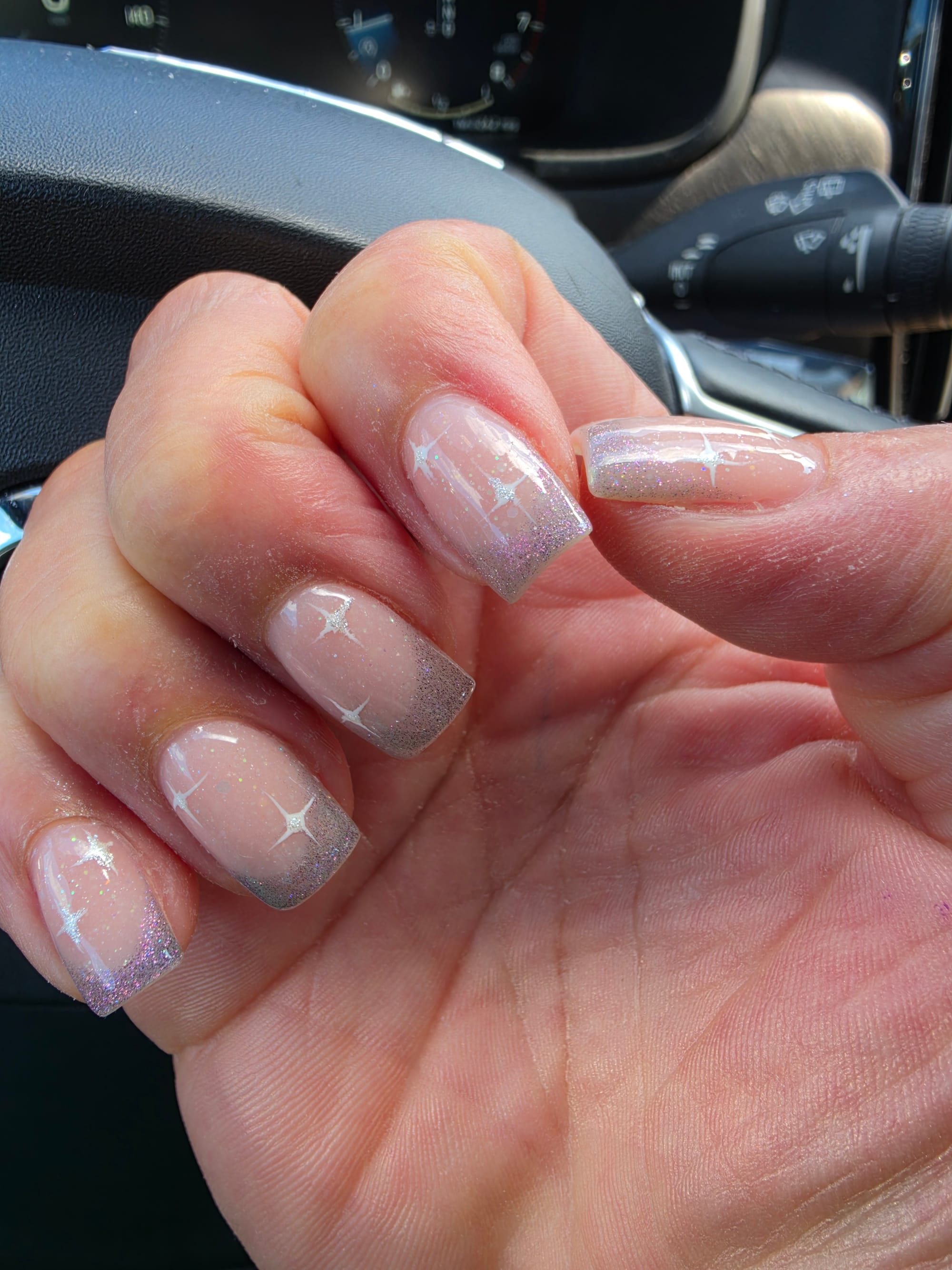 A close-up of a person'sright hand resting against a black car steering wheel, showcasing a fresh square-shaped nails with a French manicure style silver glitter gradient at the tips and celestial-themed design. of white stars.