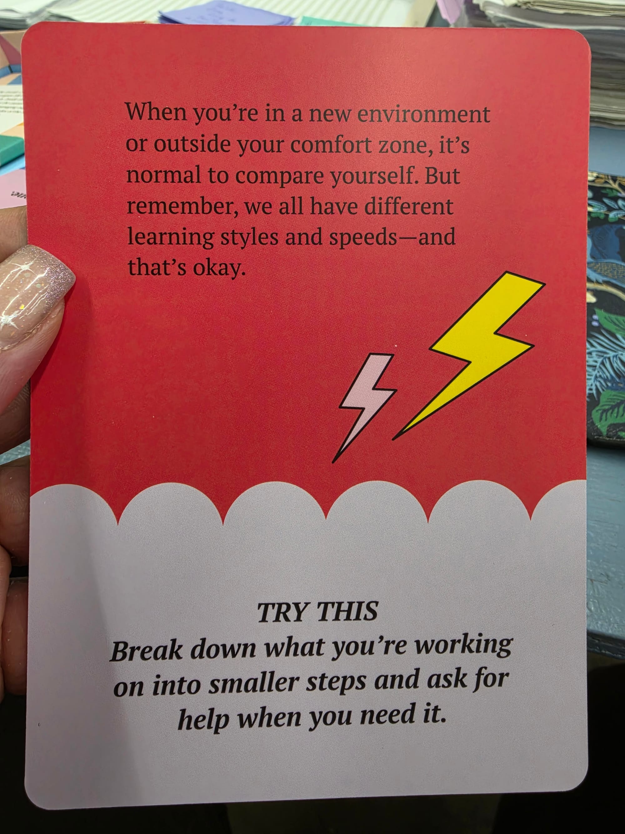 A hand holds a rectangular card with a red top half and a white bottom half, featuring text and multicolored lightning bolts. The top red section reads, "When you’re in a new environment or outside your comfort zone, it’s normal to compare yourself. But remember, we all have different learning styles and speeds—and that’s okay." The bottom white section reads: The top red section contains black text that reads, "TRY THIS" and "Break down what you’re working on into smaller steps and ask for help when you need it."