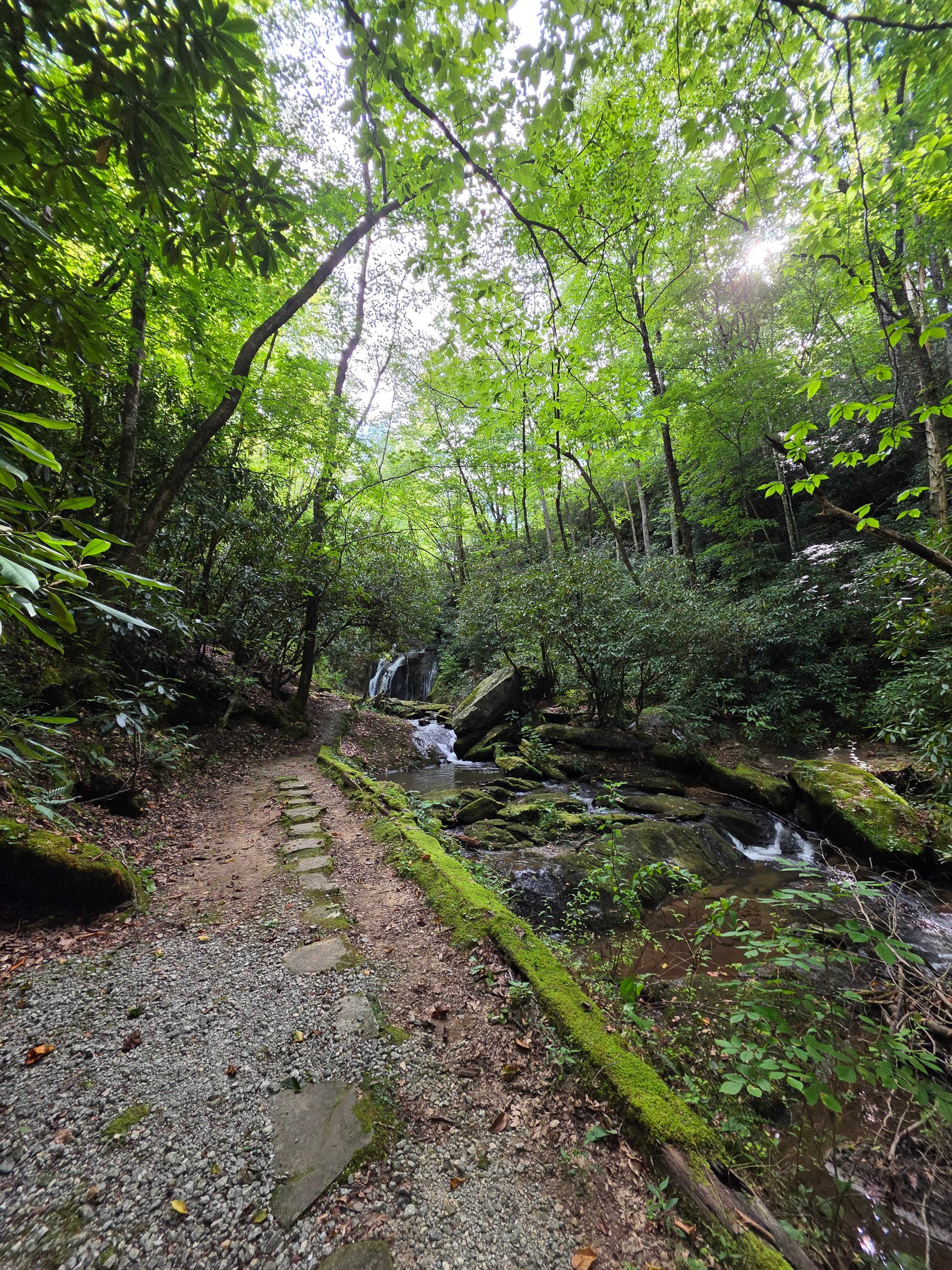 A medium-sized waterfall cascades down into a creek bed. There is a dirt and gravel trail running alongside the creek.