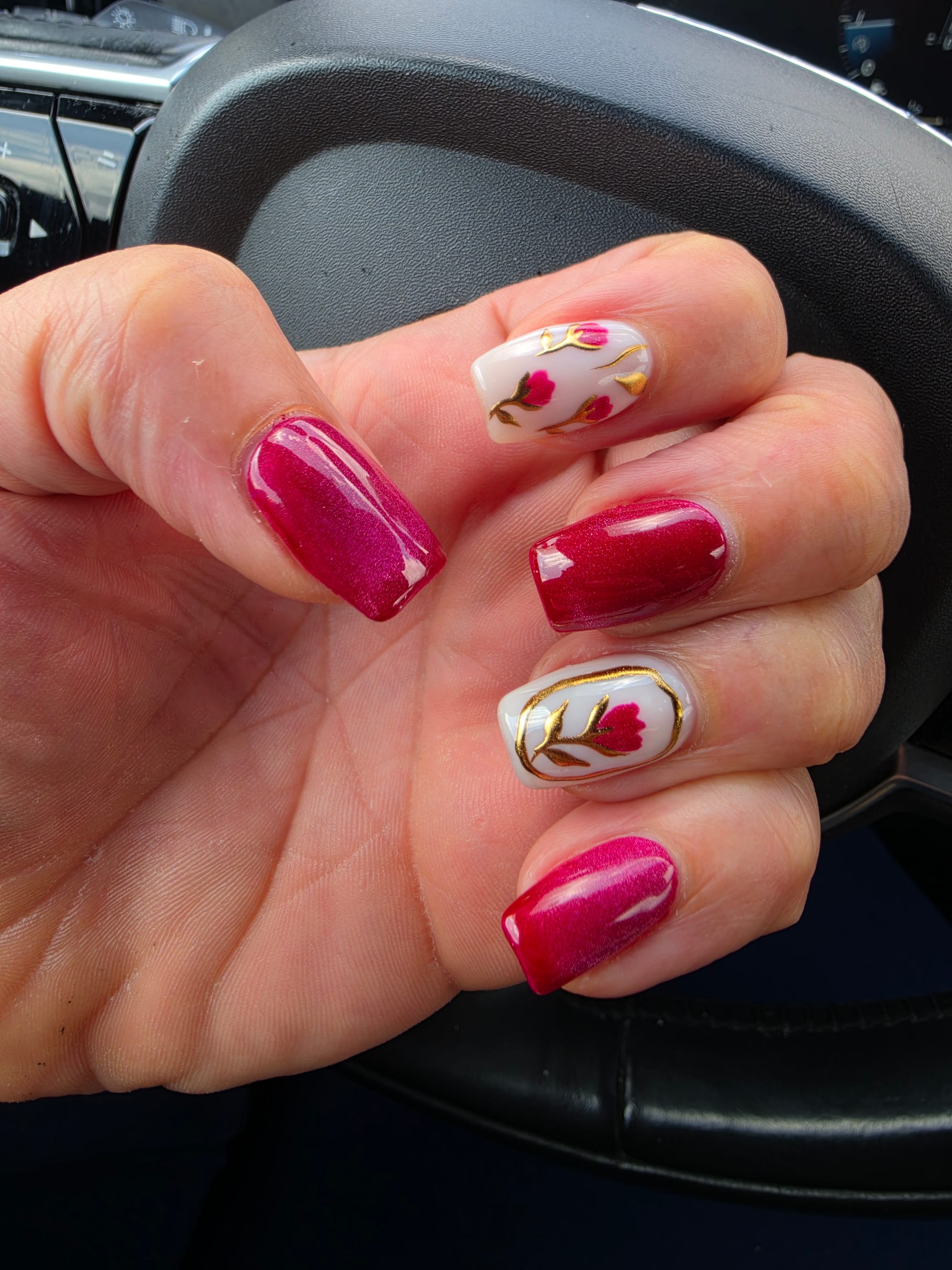 Close up of a person's left hand resting against a black steering wheel, showcasing a fresh manicure of medium-length square nails. The thumb, middle finger, and pinky sport a magenta-red nail polish with a subtle cat-eye or metallic depth. The index and ring fingers feature milky-white base with three hand-painted pink tulip buds on golden stems.