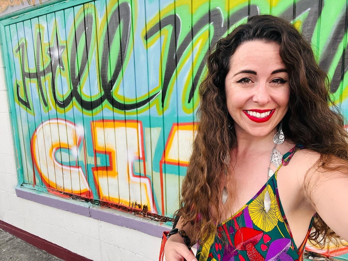 A white woman with dark brown curly hair and red lipstick, wearing a colorful mushroom-print dress takes a selfie in front of a brightly spray painted wall that reads Hillman City. 