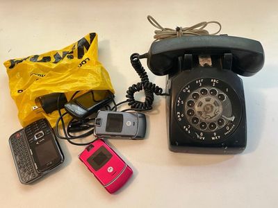 Photo of a bunch of old flip phones spilling out of a yellow NiKON bag. Beside it is a well-loved black rotary telephone. 
