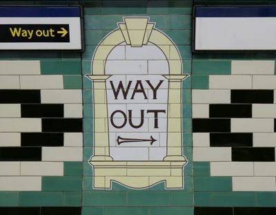 green and white and black tiles on a subway station wall, in the middle a yellow arch pattern with a white background and WAY OUT with a black arrow.