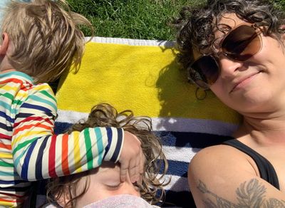 Two small children lay next to a woman on a beach towel. 