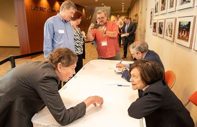 Photo of an author signing with Robert and Ina Caro on one side of the table and fans photographing Robert signing a book on the other. In the foreground, two women lean in close to talk.