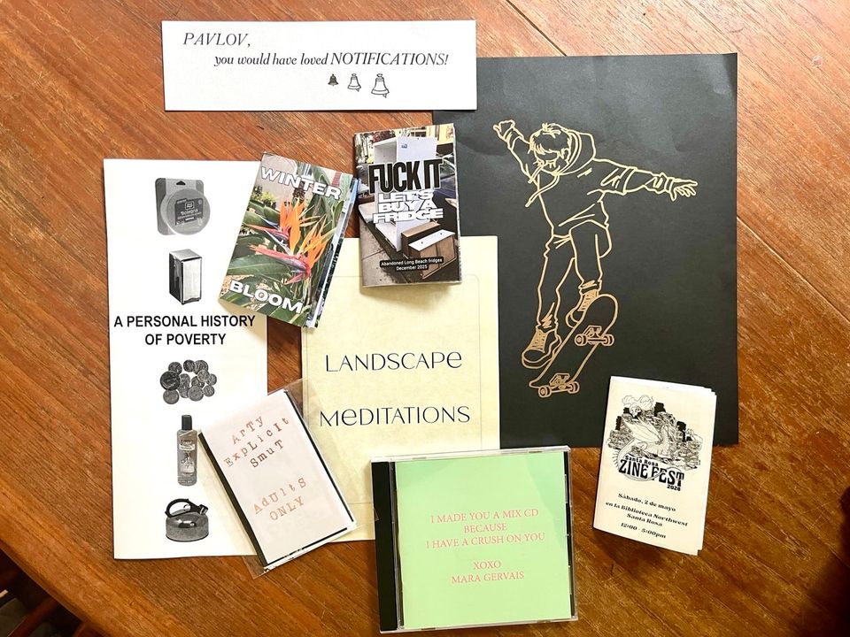 an array of zines, an art print, a cd and a bookmark laid out artfully on a brown wooden tabletop.