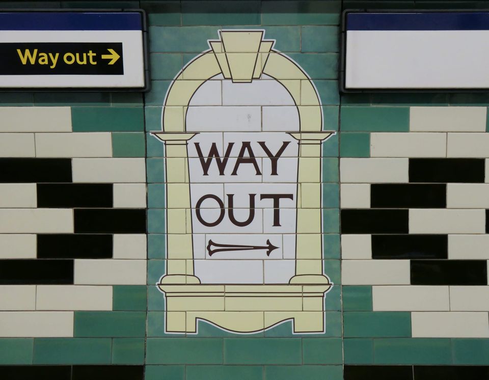 green and white and black tiles on a subway station wall, in the middle a yellow arch pattern with a white background and WAY OUT with a black arrow.