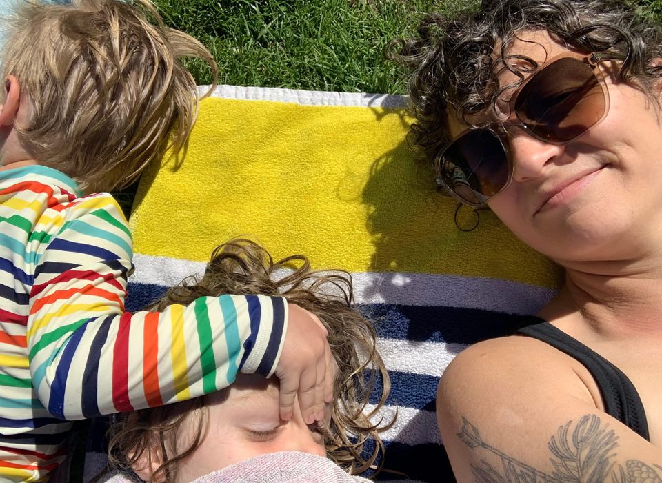 Two small children lay next to a woman on a beach towel. 