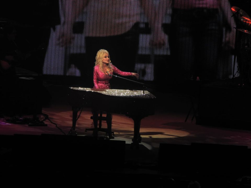 A blonde white woman in a pink rhinestone suit sits at a black grand piano on a dark stage.