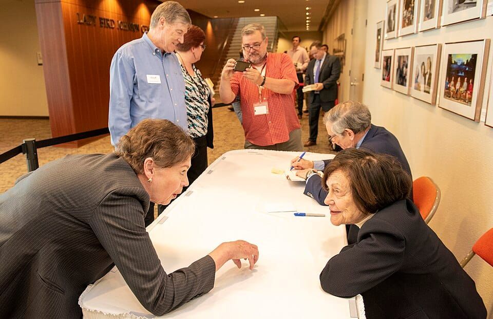 Photo of an author signing with Robert and Ina Caro on one side of the table and fans photographing Robert signing a book on the other. In the foreground, two women lean in close to talk.