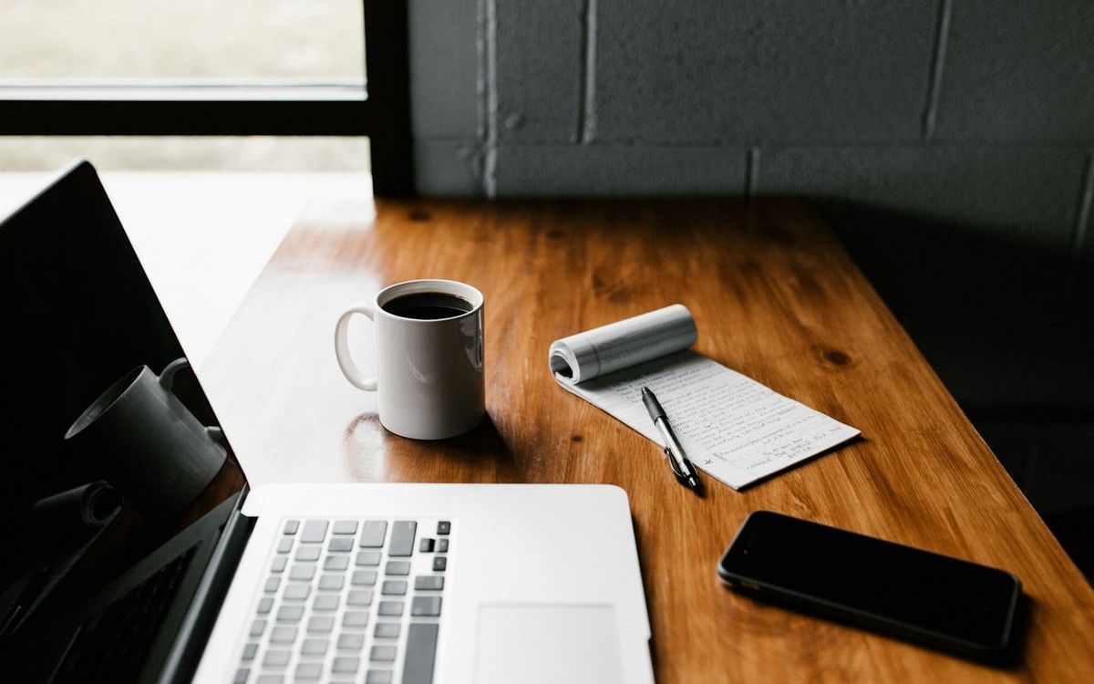 A laptop, coffee mug, phone and small notebook on a table beside a window