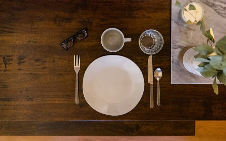 A single place setting with a plate, cutlery, glasses, and reading glasses on a wooden table.