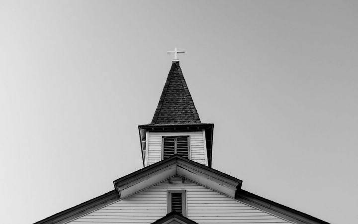 The steeple and cross of a small clapboard church in black and white