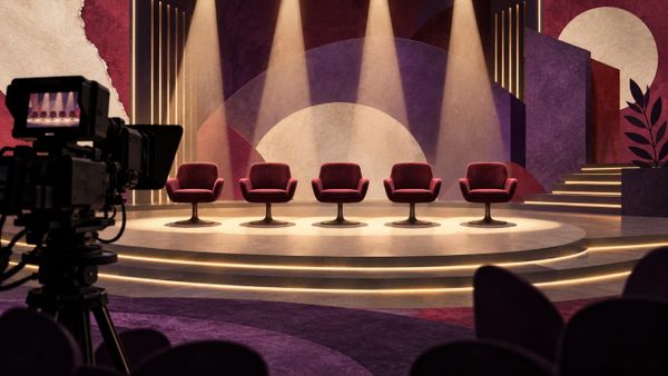 Telecamera davanti a cinque poltrone vuote illuminate in uno studio immaginario per le nomination GF VIP, senza concorrenti o loghi.