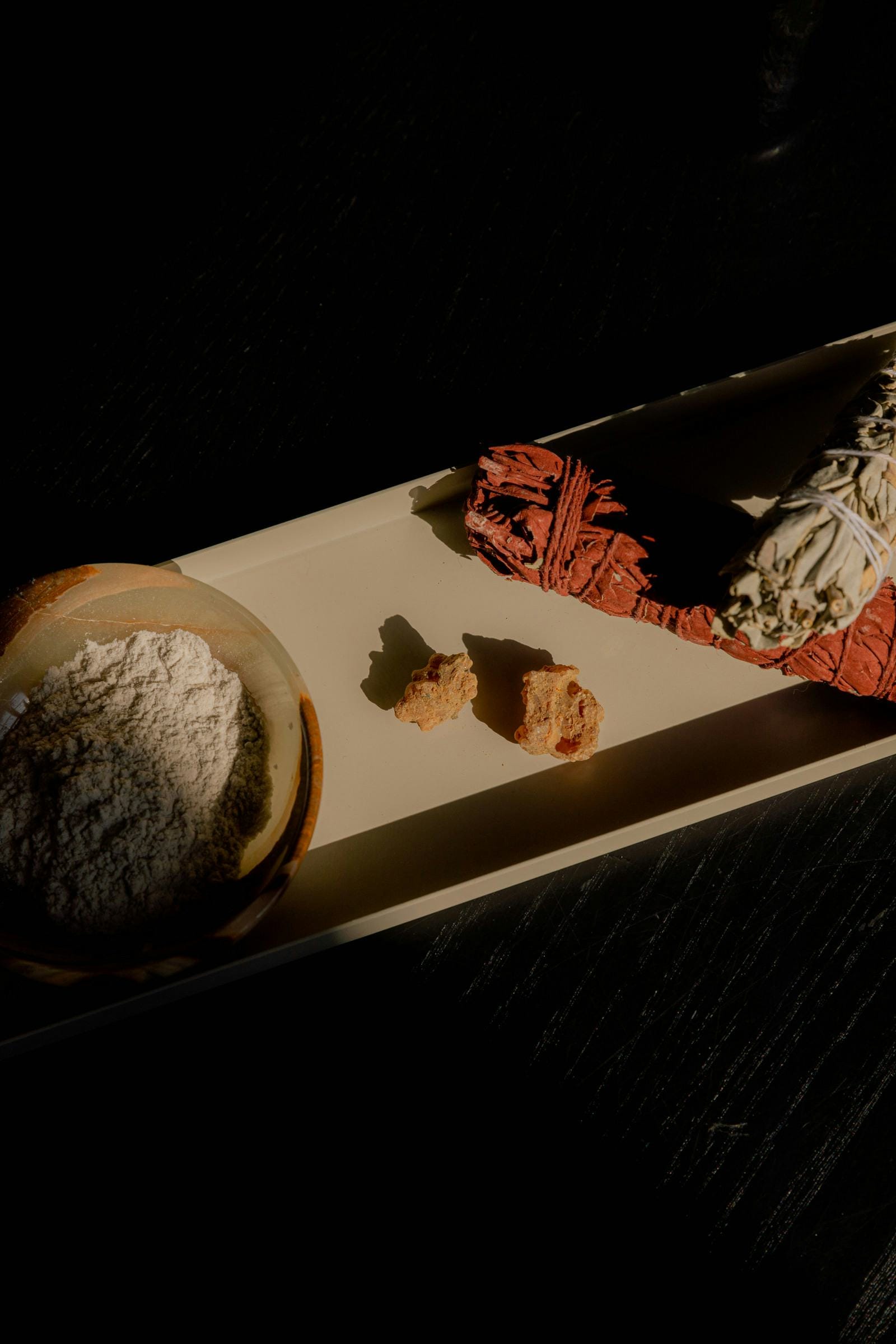 Smudge bundles and raw resin on a tray, lit by a single shaft of low light against near-black