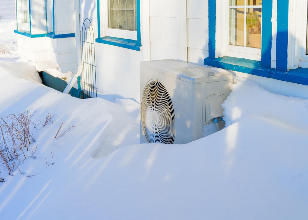 A stock image of a heat pump in deep snow outside a house.