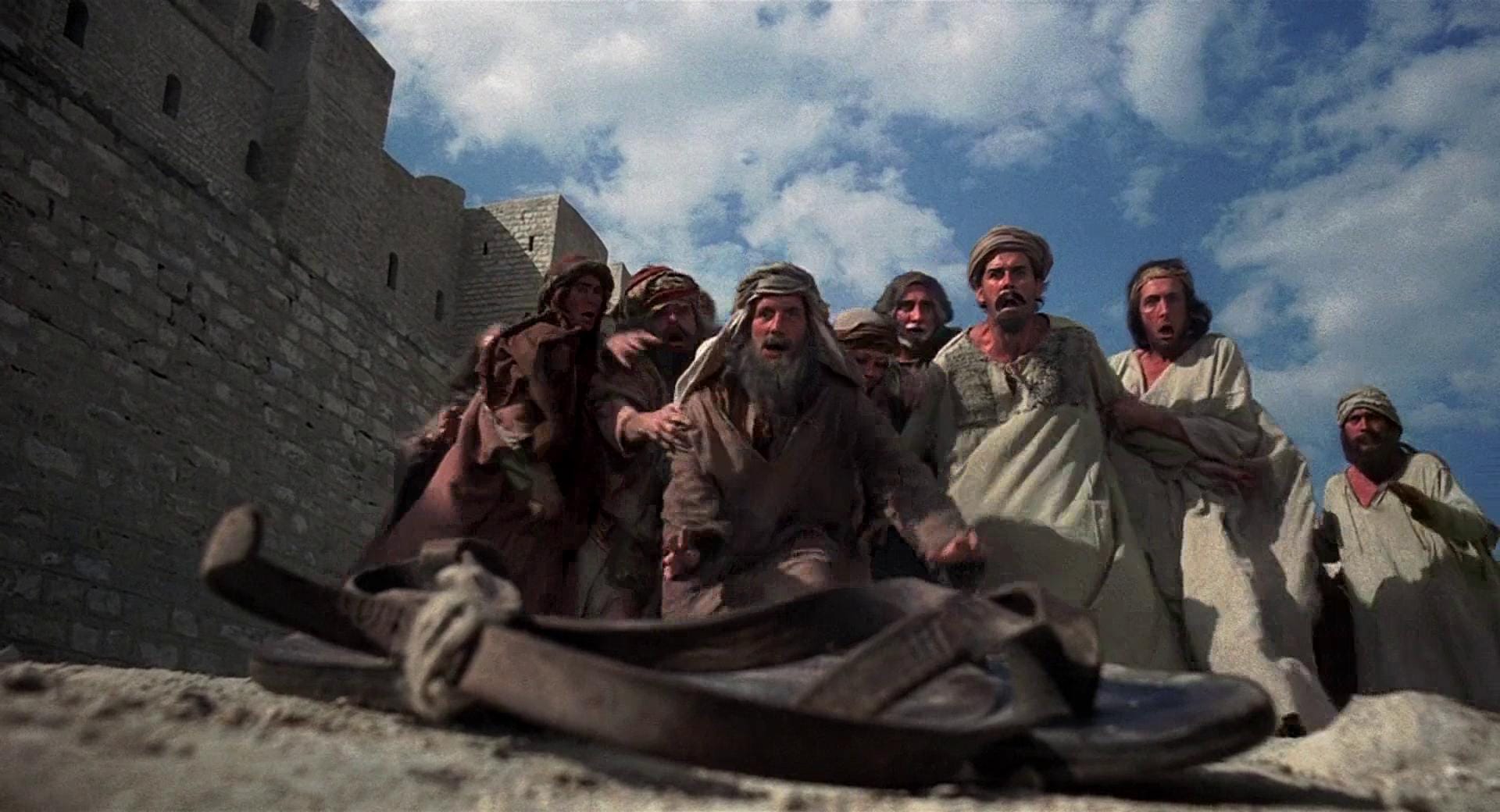 A screencap from the film Life of Brian, with Michael Palin, John Cleese and colleagues staring in wonder at an abandoned sandal.