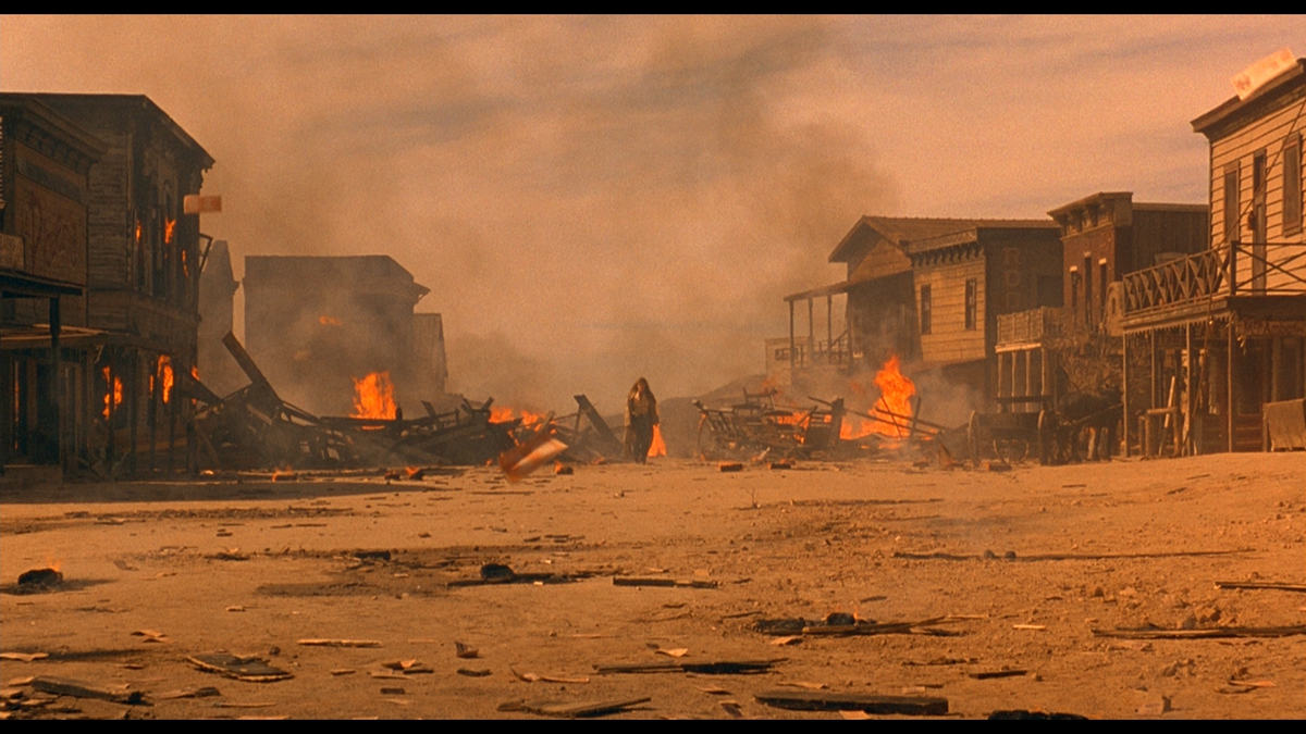 A lone figure stands among burning buildings in a dusty Old West town.
