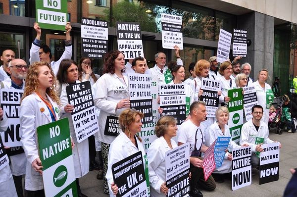 A group of about two dozen people of different ages in white lab coats hold protest signs outside a building.