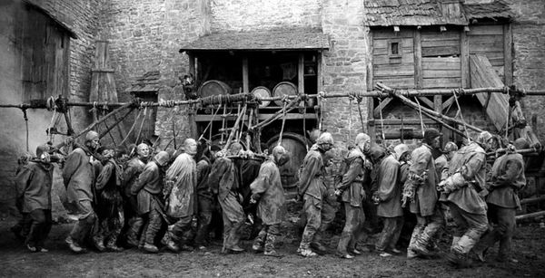 A black and white still from a movie, in which figures dressed in filthy rags are walking in single file, their necks tethered to a poles suspended above.