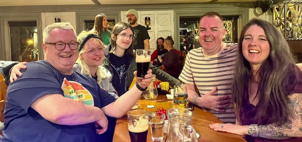 A group of people sit smiling as one holds a pint of guinness with a successfully split G