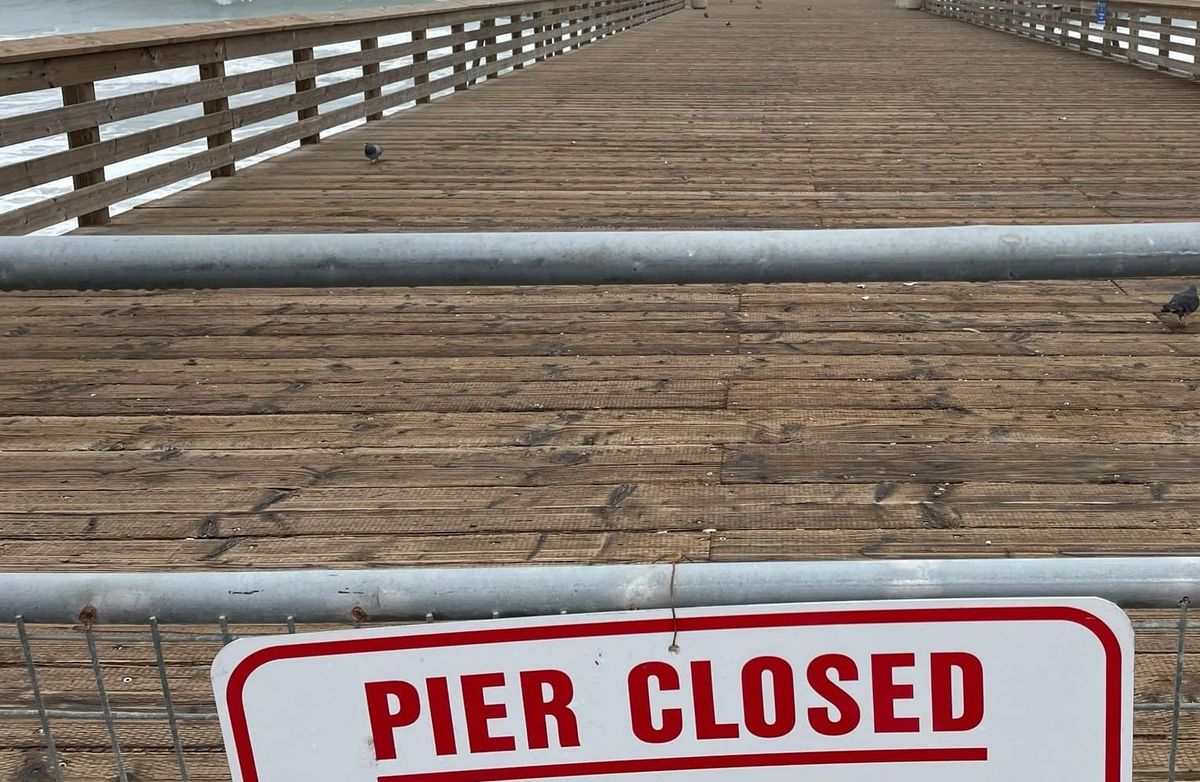 A 'Pier Closed' sign stands at the entrance to a wooden walkway overlooking a restless ocean.