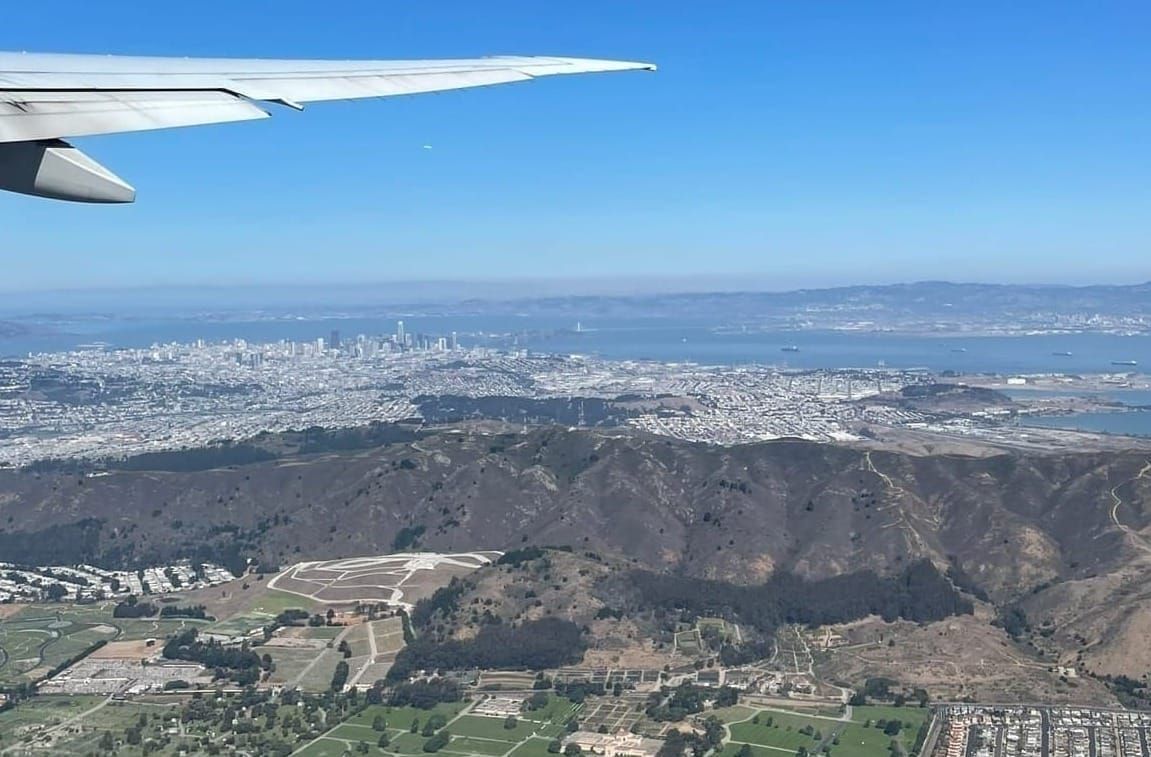 An aerial bird’s-eye view from an airplane window showing a city nestled between a mountain range and a large lake during takeoff.