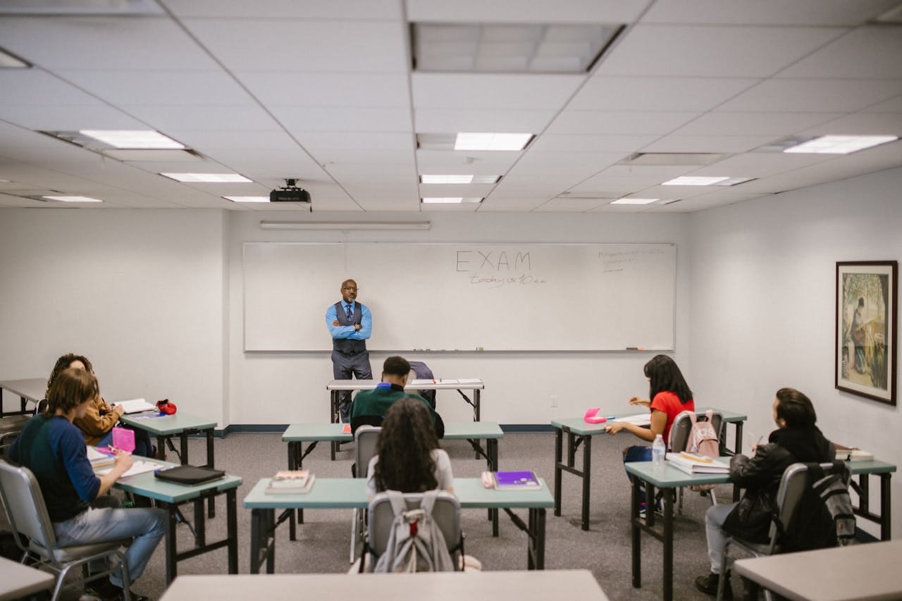 Teacher standing at the front of a classroom with 'EXAM' written on the whiteboard, students seated at individual desks