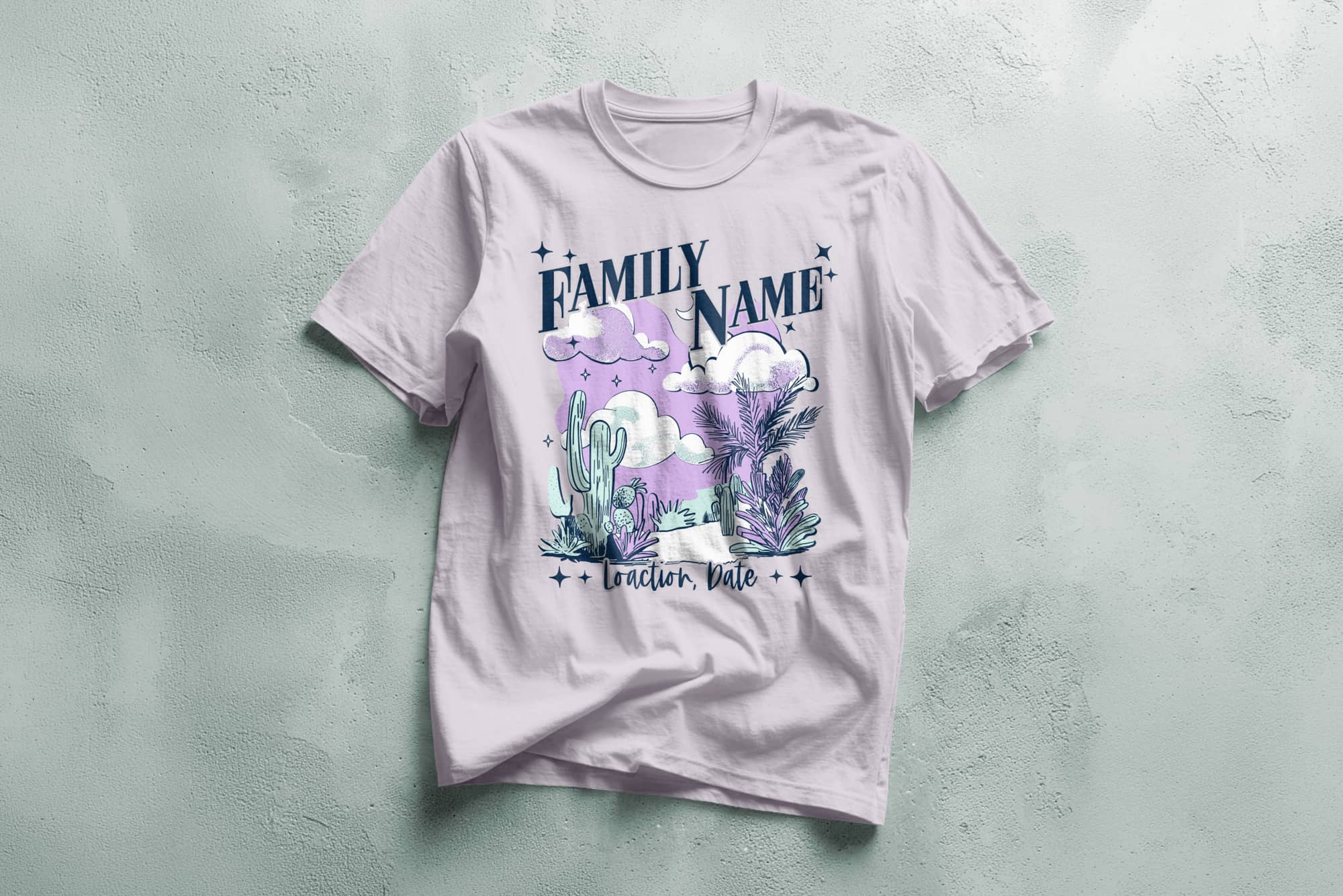 A lavender T-shirt with a desert illustration featuring clouds, cacti, and stars. "Family Name" is at the top, with "Location, Date" below in elegant script.