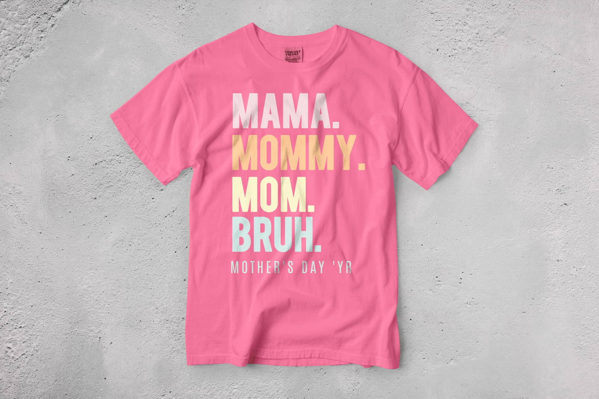 Pink T-shirt with text: "MAMA. MOMMY. MOM. BRUH." in white, yellow, and blue letters. The bottom reads "MOTHER'S DAY YR" in white. Playful tone.
