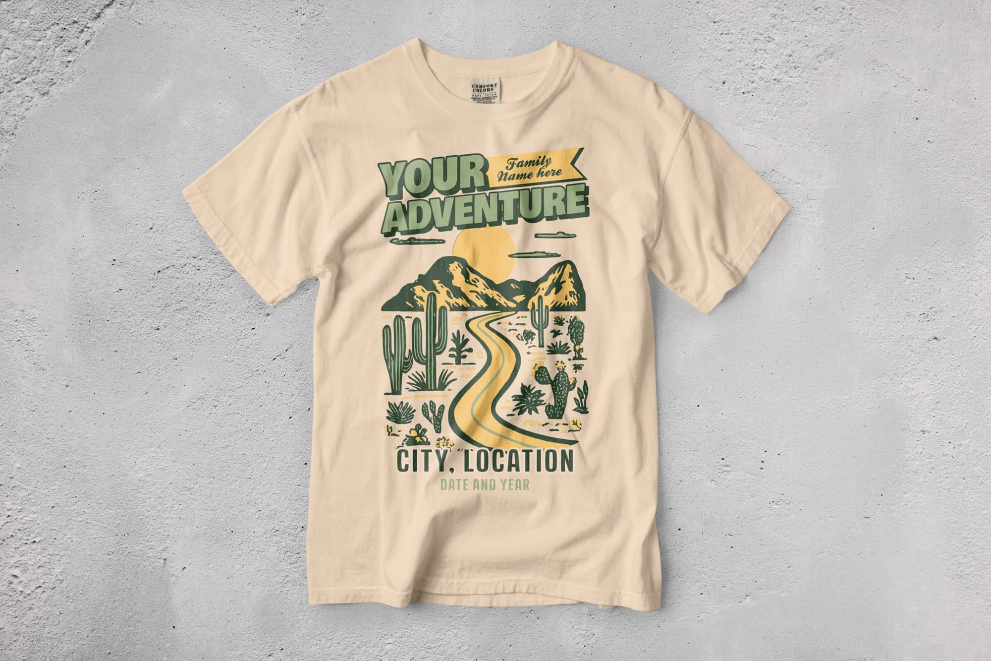 Beige T-shirt on concrete, featuring a graphic of a winding road through cacti and mountains under a sunrise. Text: "Your Adventure" with customizable details.