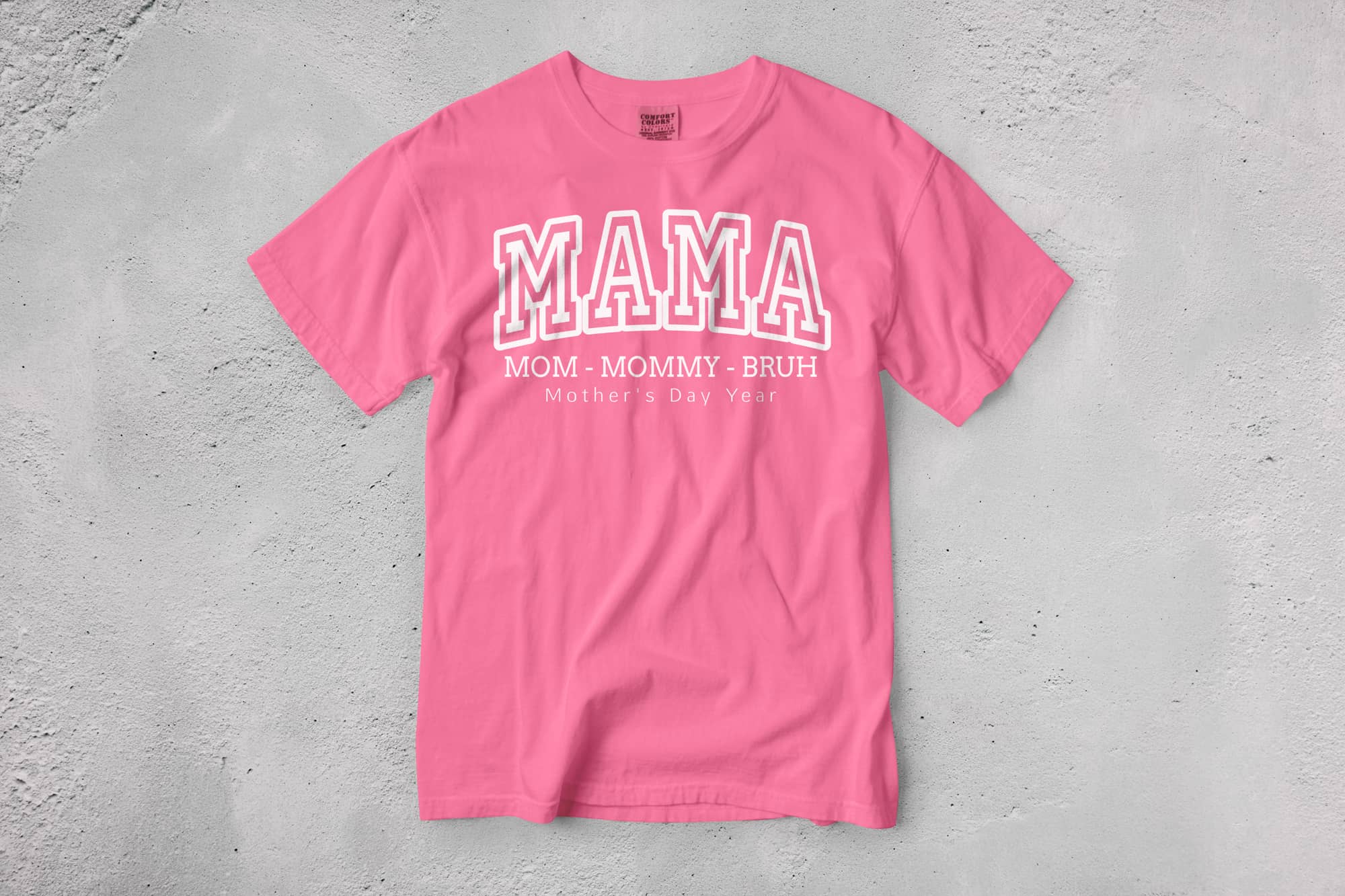 A pink T-shirt with bold, white text reading "MAMA" above smaller text: "Mom - Mommy - Bruh." It conveys a playful, affectionate tone.