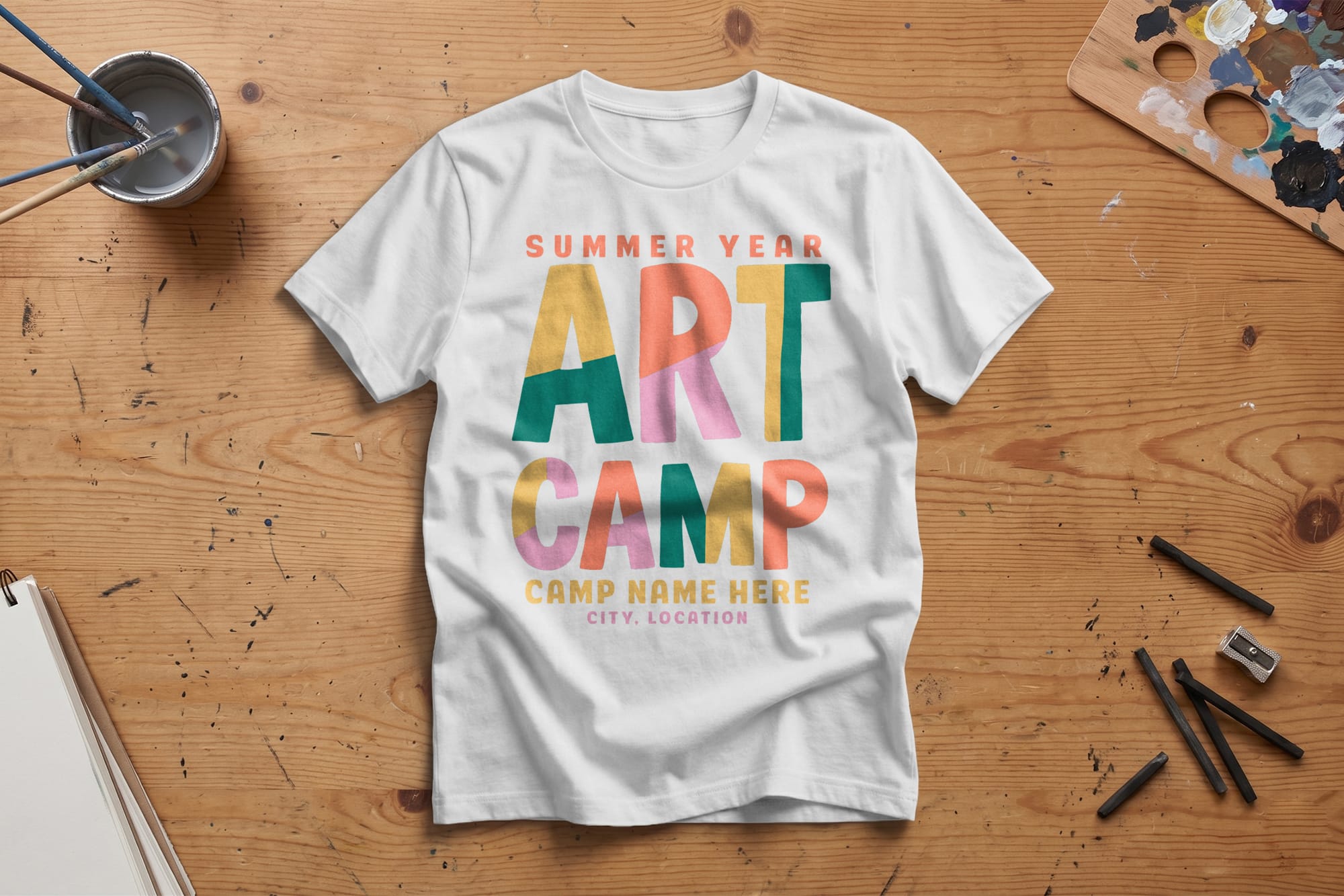 White T-shirt on a wooden table reads “Summer Year Art Camp” in colorful, bold letters. Nearby are black pencils, paint palette, and a cup of water.