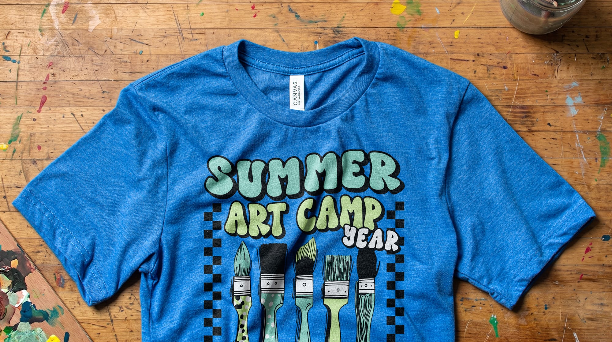 Blue T-shirt laid flat on a paint-splattered wooden table. The shirt features "Summer Art Camp" text and five paintbrushes illustrated below.