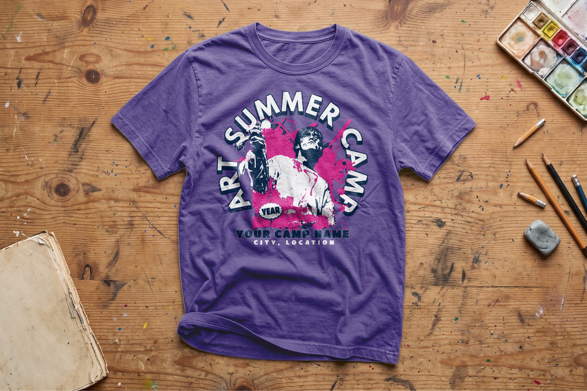 Purple T-shirt with an “Art Summer Camp” graphic showing an artist holding a paintbrush. Set on a wooden table with art supplies, conveying creativity.