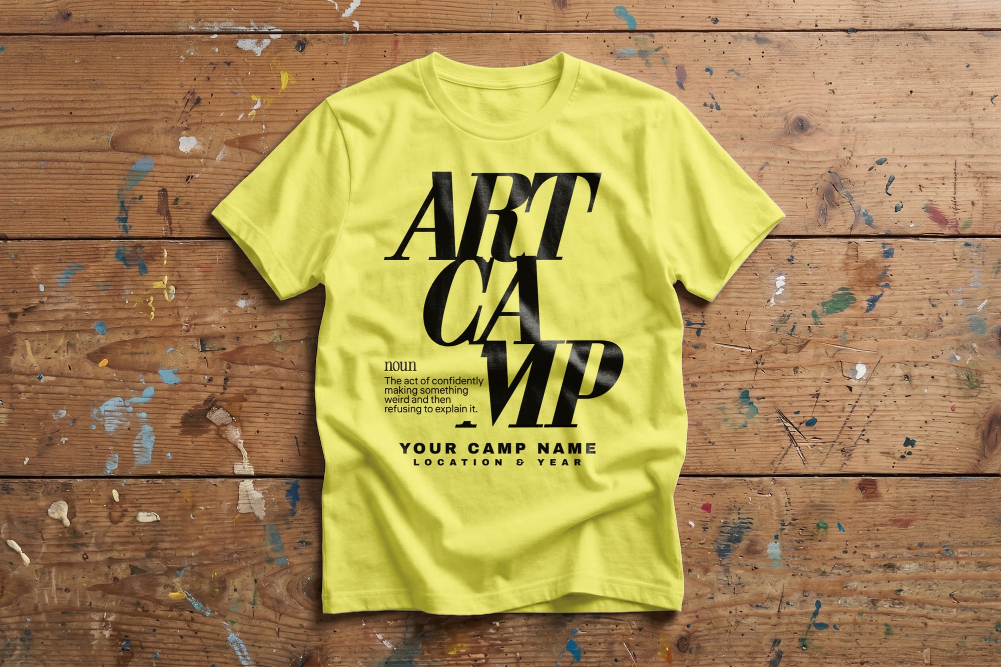 Yellow "Art Camp" T-shirt with bold black text on a wooden surface, paint splatters around. Text reads "Art Camp" with space for camp details. Bright and creative.
