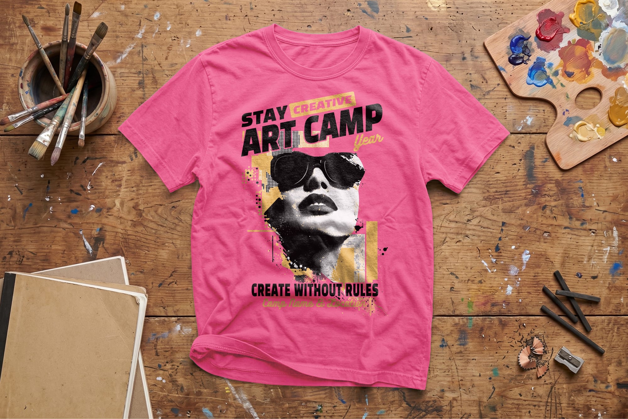 Pink T-shirt on a wooden table featuring bold text "Stay Creative Art Camp," a stylized face in sunglasses, surrounded by paint and brushes, evoking creativity.