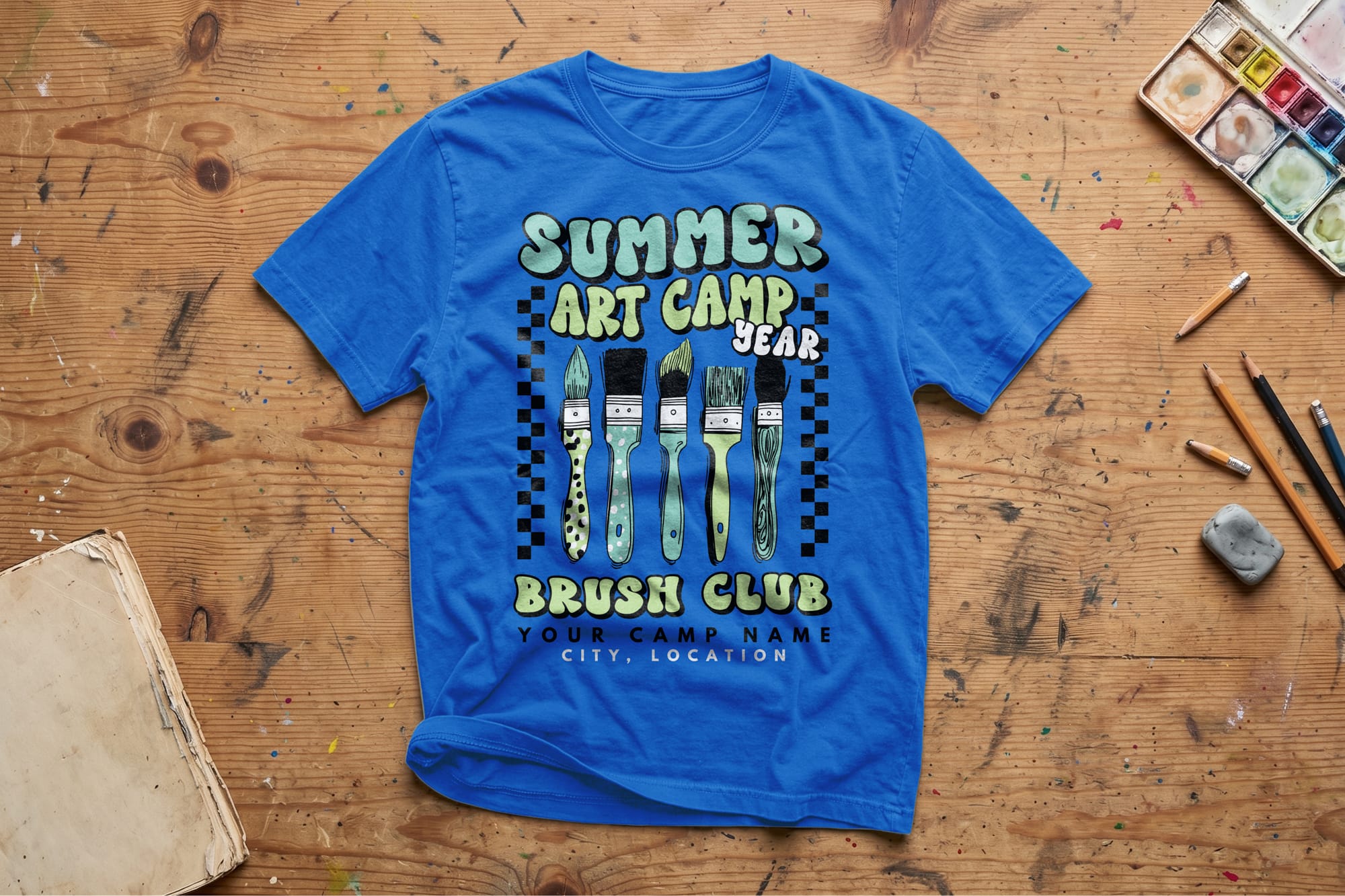 Blue T-shirt on a wooden table featuring "Summer Art Camp" with paintbrush designs. Surrounded by art supplies, it evokes creativity and fun.