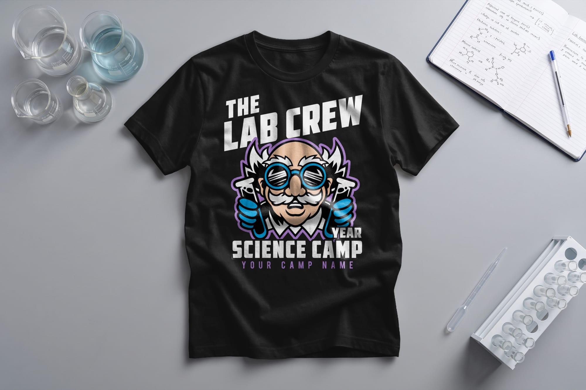 Black t-shirt with a cartoon scientist holding flasks, labeled "The Lab Crew," "Science Camp," and placeholders for year and camp name. Nearby are beakers, open notebook, pen, and test tubes, suggesting a lab setting.