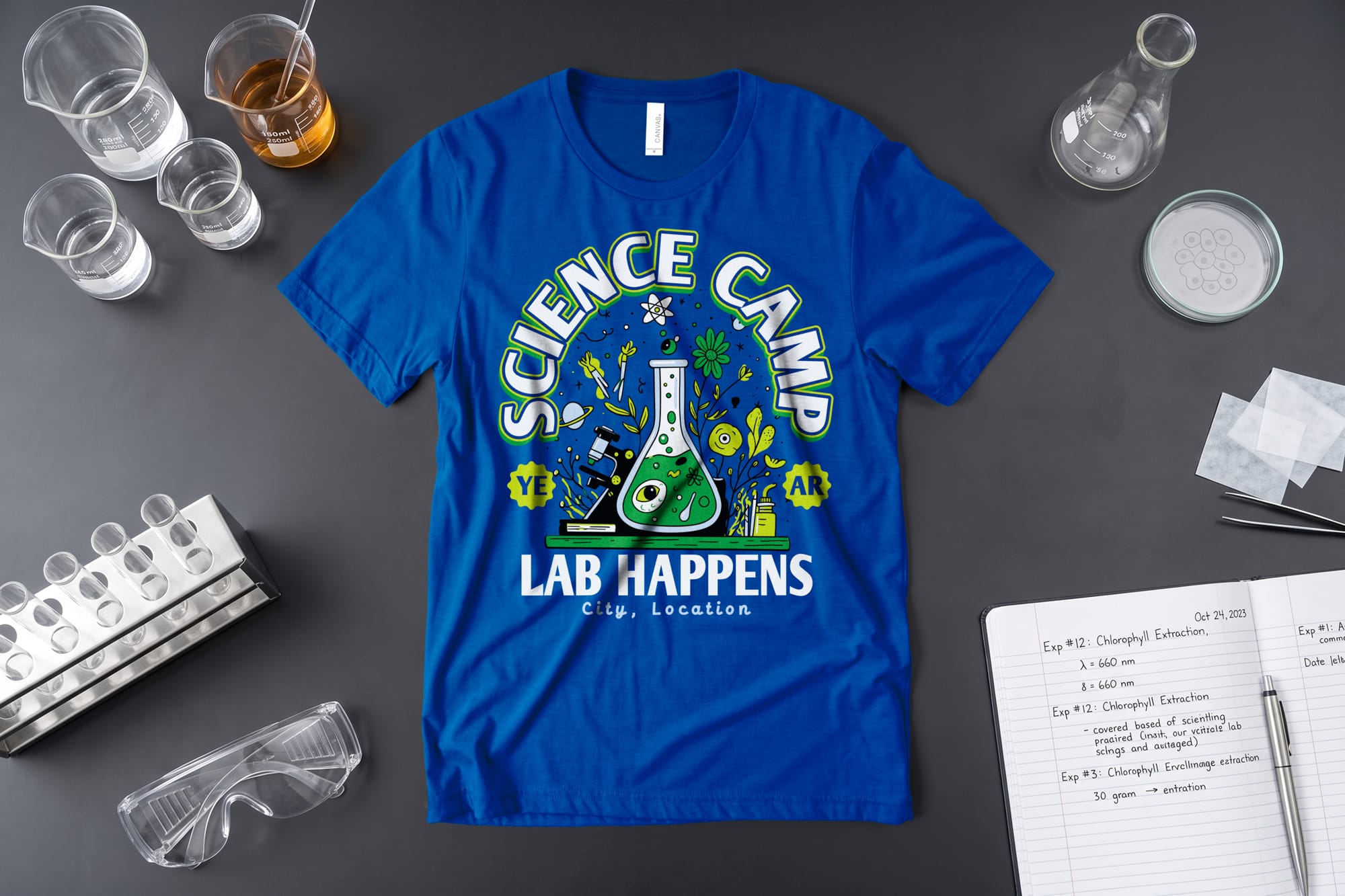 Blue T-shirt with "Science Camp, Lab Happens" text and a beaker illustration surrounded by scientific icons. Lab equipment and goggles nearby.