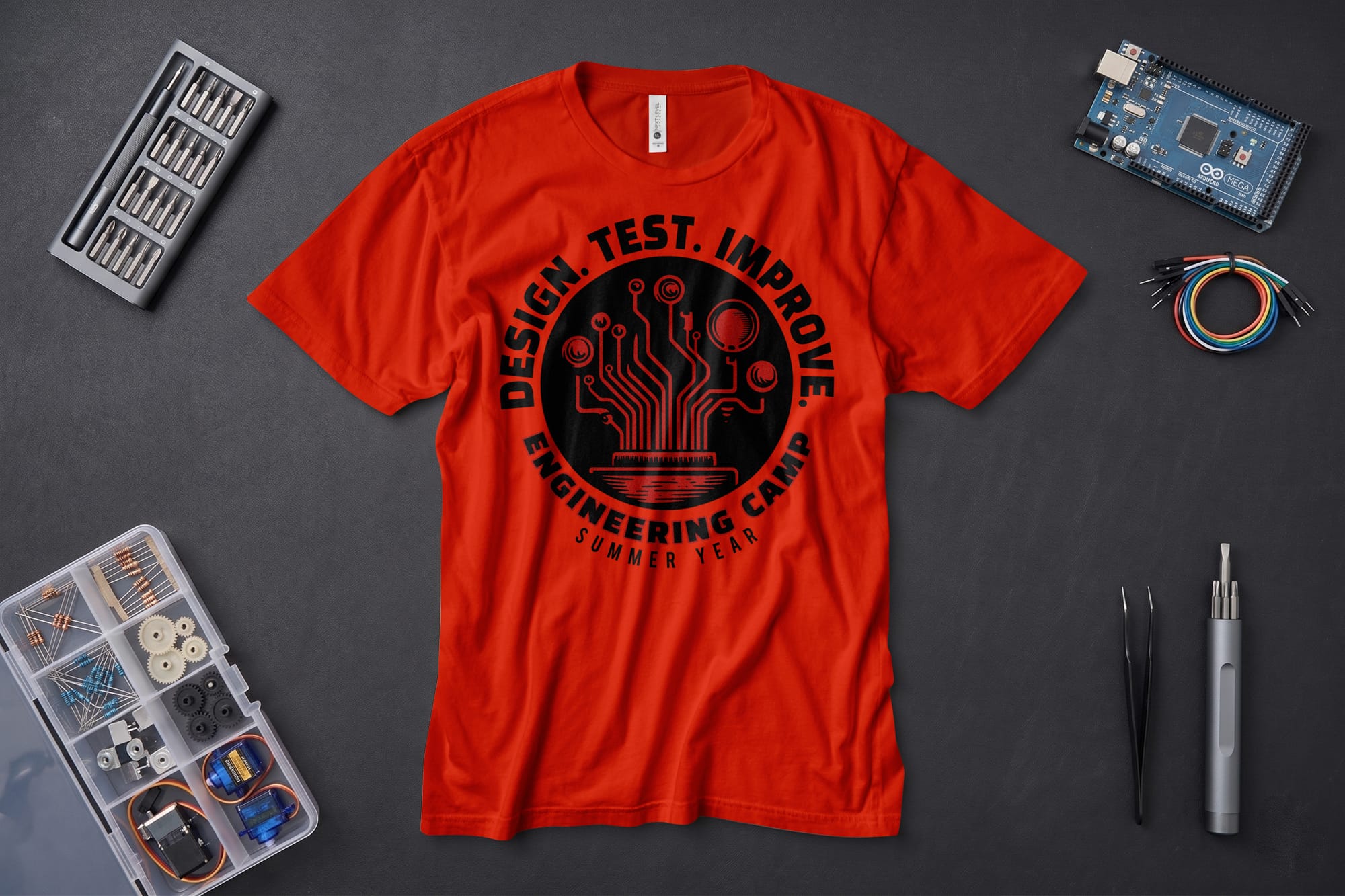 Red T-shirt with “Design. Test. Improve. Engineering Camp” text and circuit design. Surrounding it are tools, wires, microcontroller, and components.